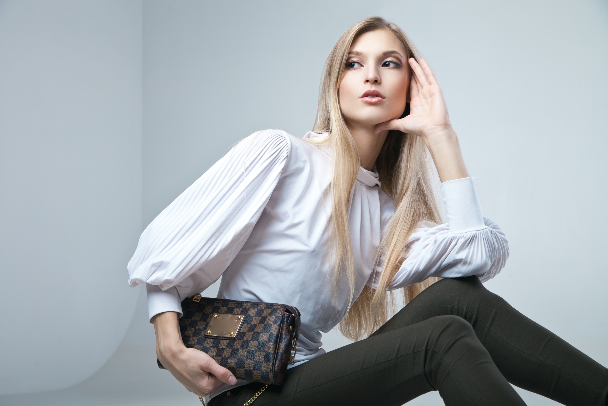 Full length casual young fashionable woman sitting with a bag in light background
